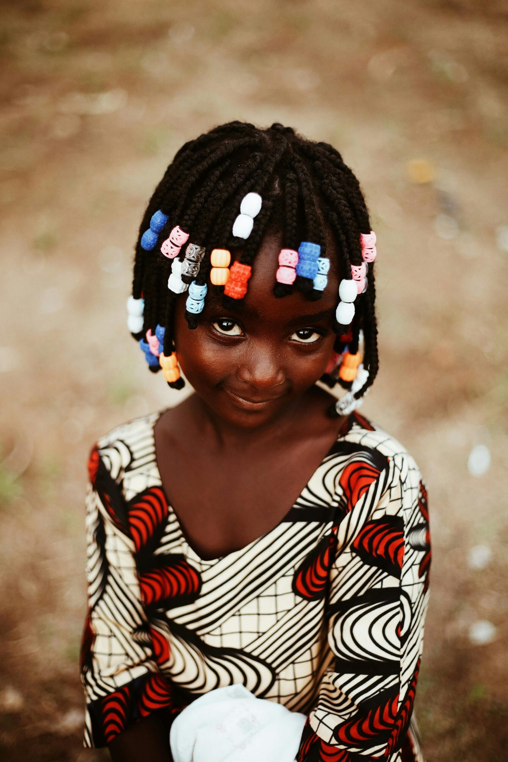 Adorable young girl in a colorful African dress with braided hair, smiling outdoors.